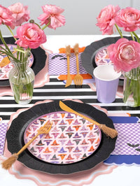 Top-down detail of two witch hat plate settings on black chargers with purple checkered napkins, gold flatware, and pink roses.