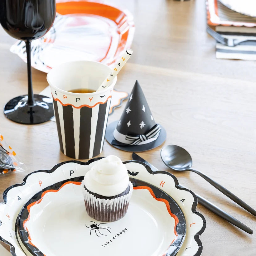 Black witch hat treat box with white stars and a black and white striped bow at a Halloween place setting next to a striped cup, cupcake and black flatware