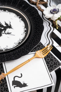 Close up angled view of a vintage witch plate and a black cat paper napkin tucked neatly under gold flatware on a striped runner.