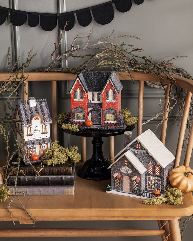 Witch Bootique raised on stacked books beside two Halloween village houses with mossy branches and a black scalloped banner