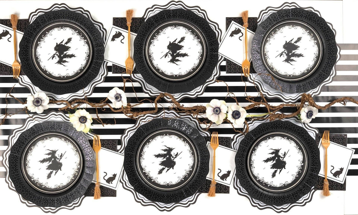 Symmetrical Halloween tablescape showing six witch silhouette plates arranged evenly along a black striped paper runner.