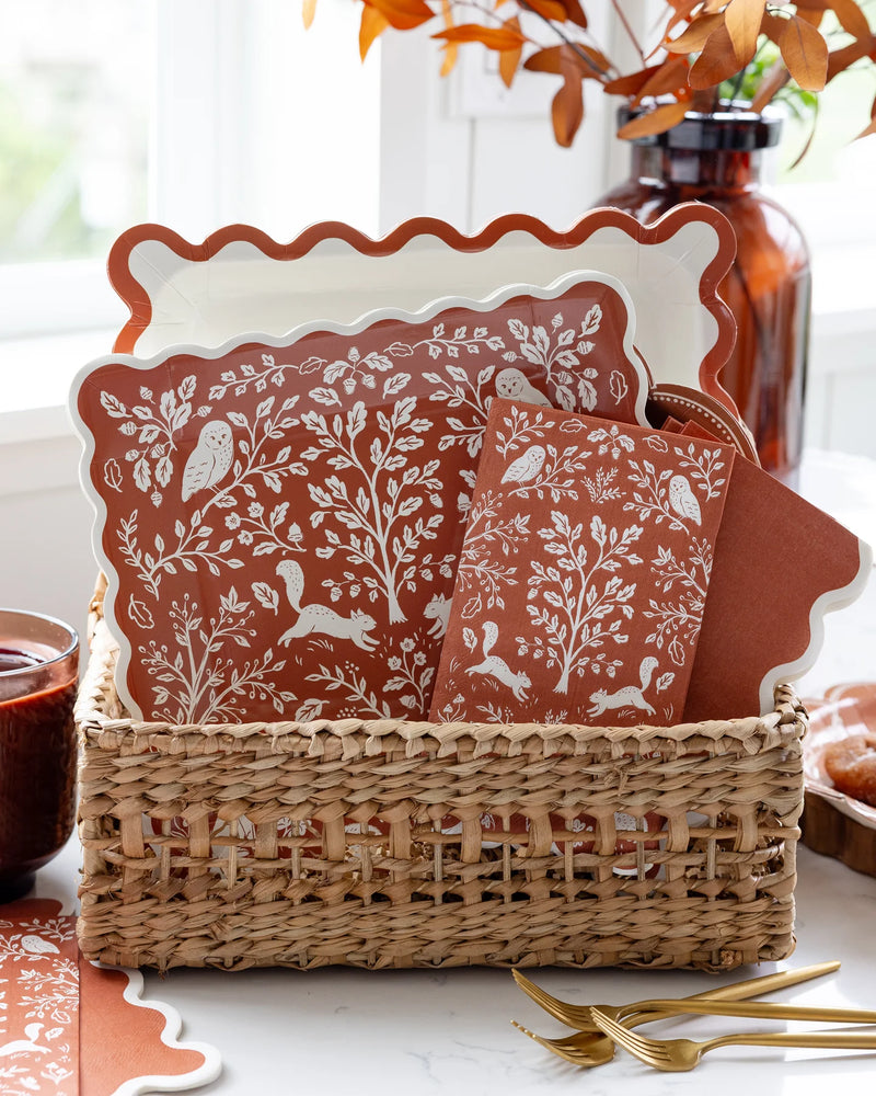 Woodland plates displayed in a woven basket — the cream ric-rac plate alongside the illustrated Acorn Grove plates, with a "Give Thanks" sign and oversized hanging autumn leaves.