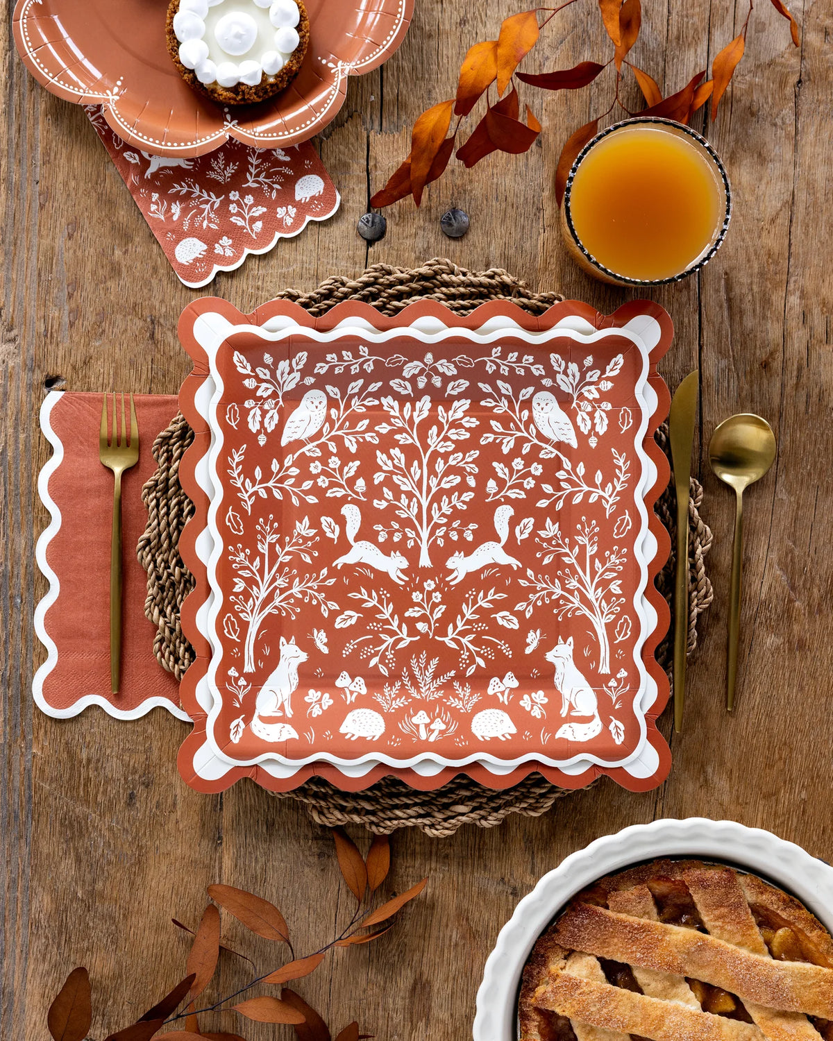 Overhead Thanksgiving place setting with a woodland print plate layered on a ric rac plate, rust napkin, gold utensils, cider, and a pie