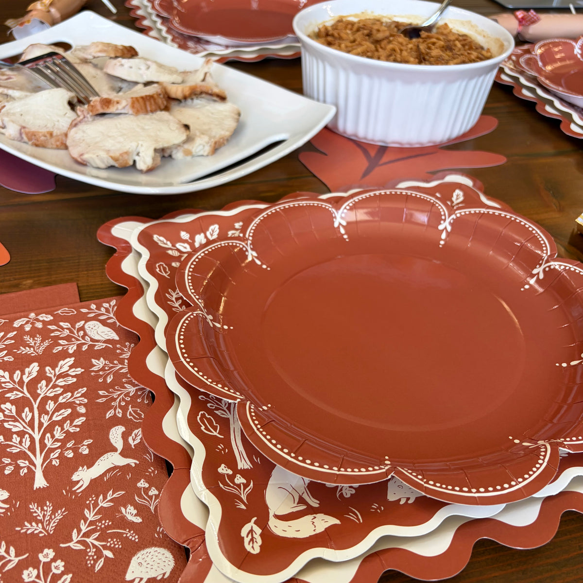 Rust scalloped plate layered on woodland and ric rac plates at a Thanksgiving dinner table with sliced turkey and a casserole