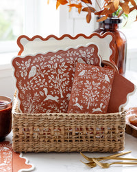 Woodland Thanksgiving plates and napkins standing in a woven basket next to gold forks and a vase of orange leaves