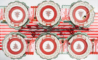 Overhead holiday table with multiple wreath-plate settings, tree plates, a striped runner, red ornaments, and gold cutlery.
