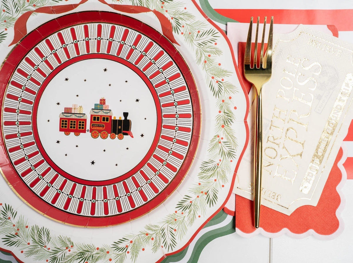 Close-up of a North Pole Express toy-train plate on the wreath dinner plate and green charger, with a gold fork and a striped runner.