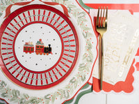 Close-up of a North Pole Express toy-train plate on the wreath dinner plate and green charger, with a gold fork and a striped runner.